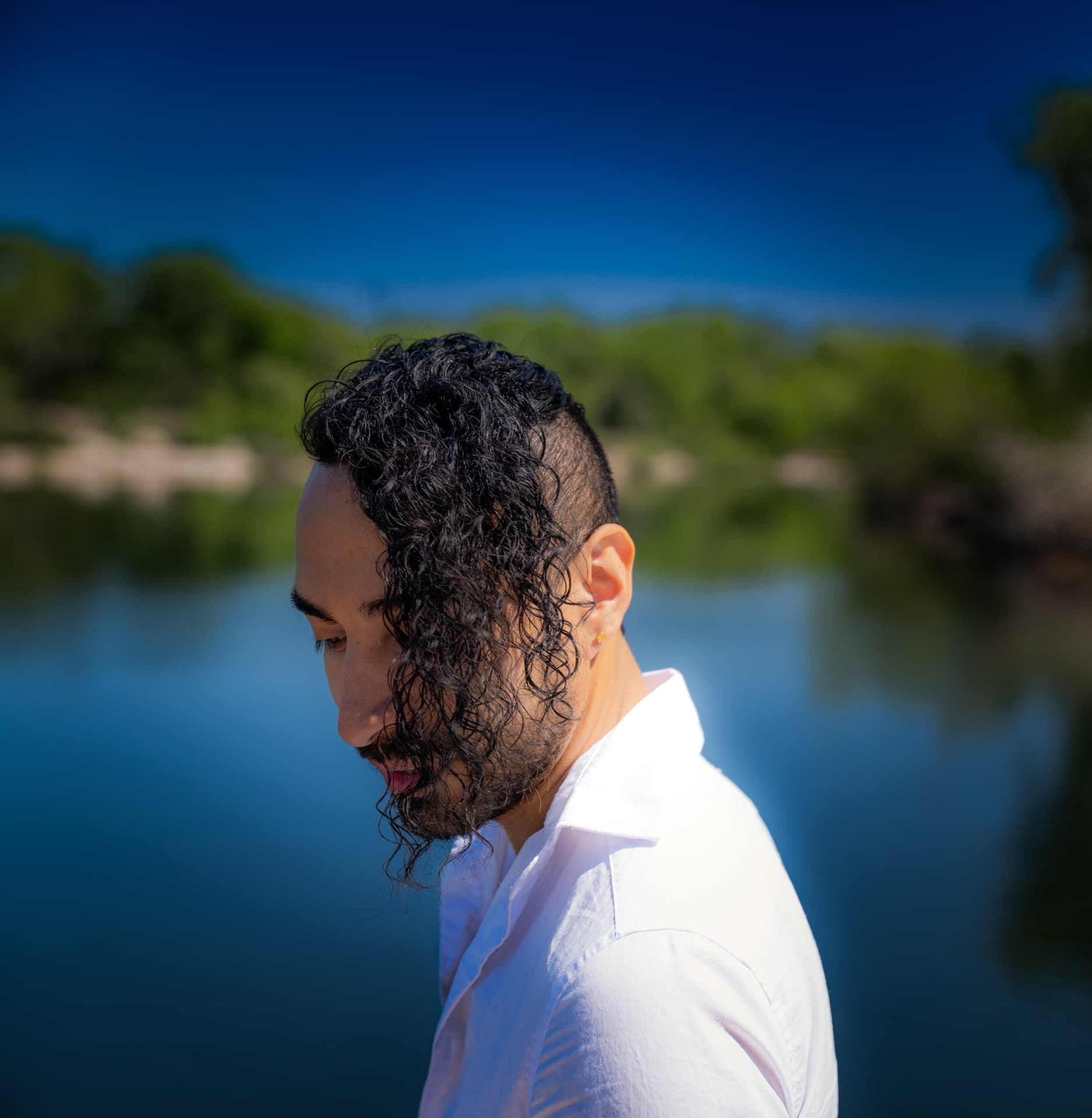 Man with curly dark hair in a white shirt beside a bright blue lake